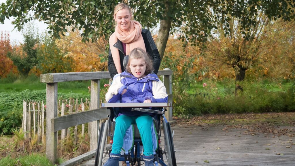 A young girl is sitting in a wheelchair and a female support worker is helping her move around while strolling in a park