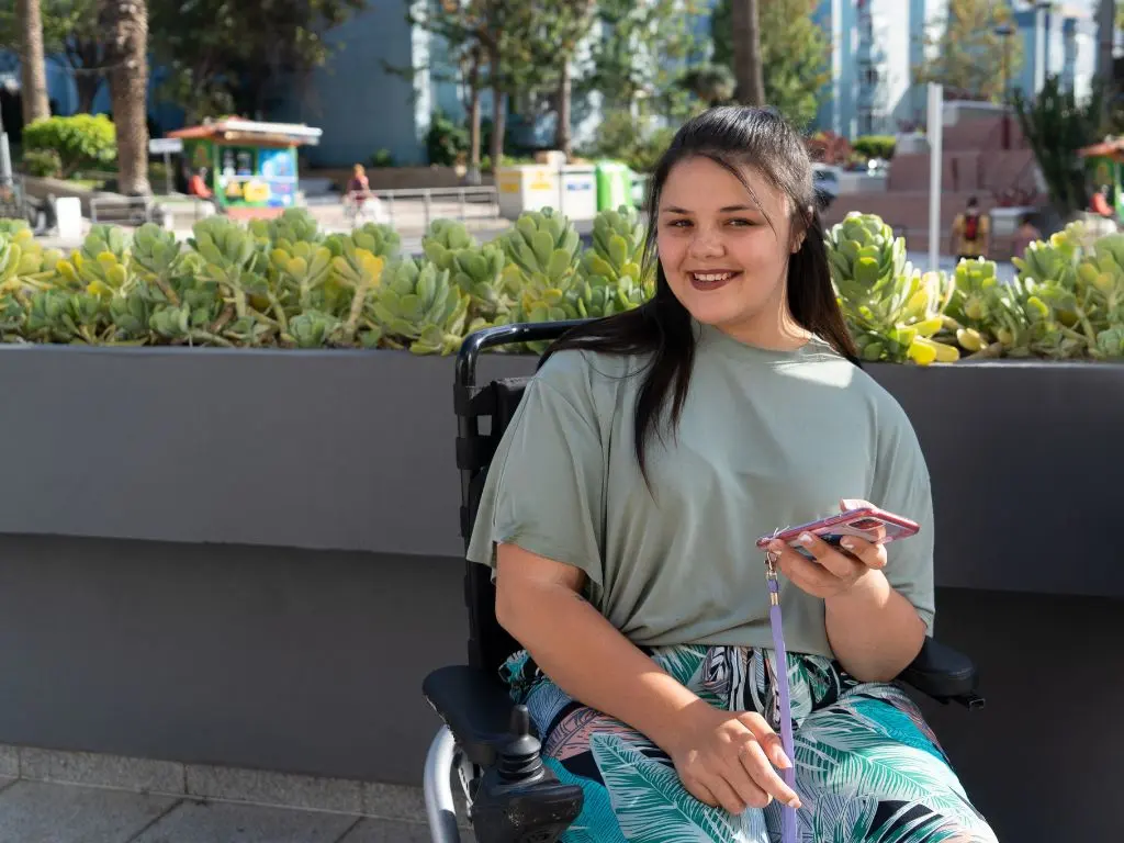 A young woman, in her wheelchair, holding her phone and smiling